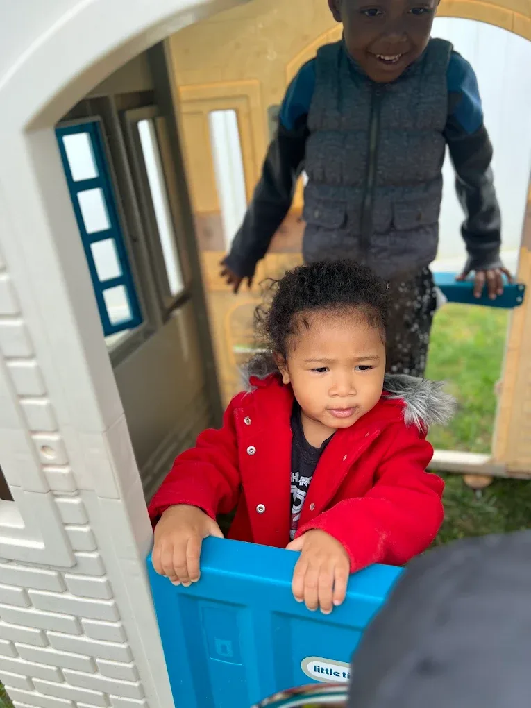 Toddler in a red coat smiling in the playhouse with a friend